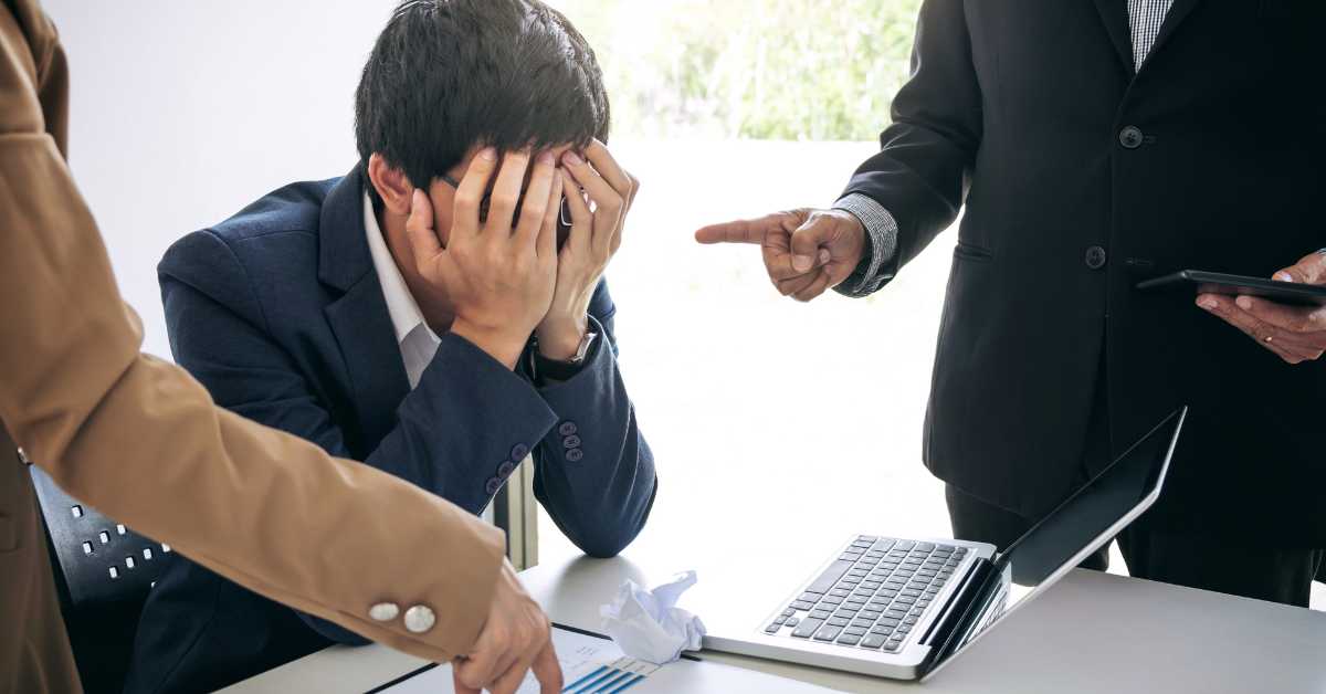A man in a suit sits at a desk with his head in his hands, showing signs of workplace burnout as two colleagues gesture and point at documents and a laptop in front of him.