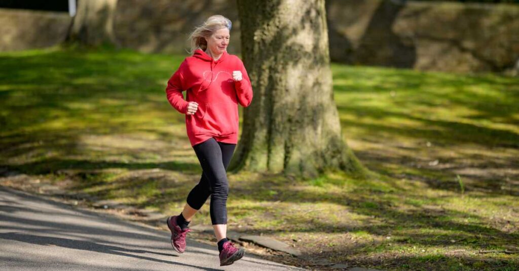 An older woman wearing a red hoodie and black leggings jogs on a paved path in a sunny park, finding relief from workplace burnout amid the trees and grass.