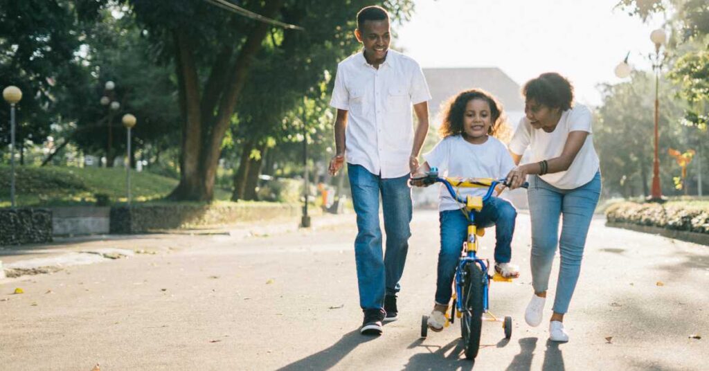 A child rides a bicycle while two adults walk beside, supporting and encouraging them on a sunny day in the park—just like strong support can help overcome job burnout and regain balance in life.