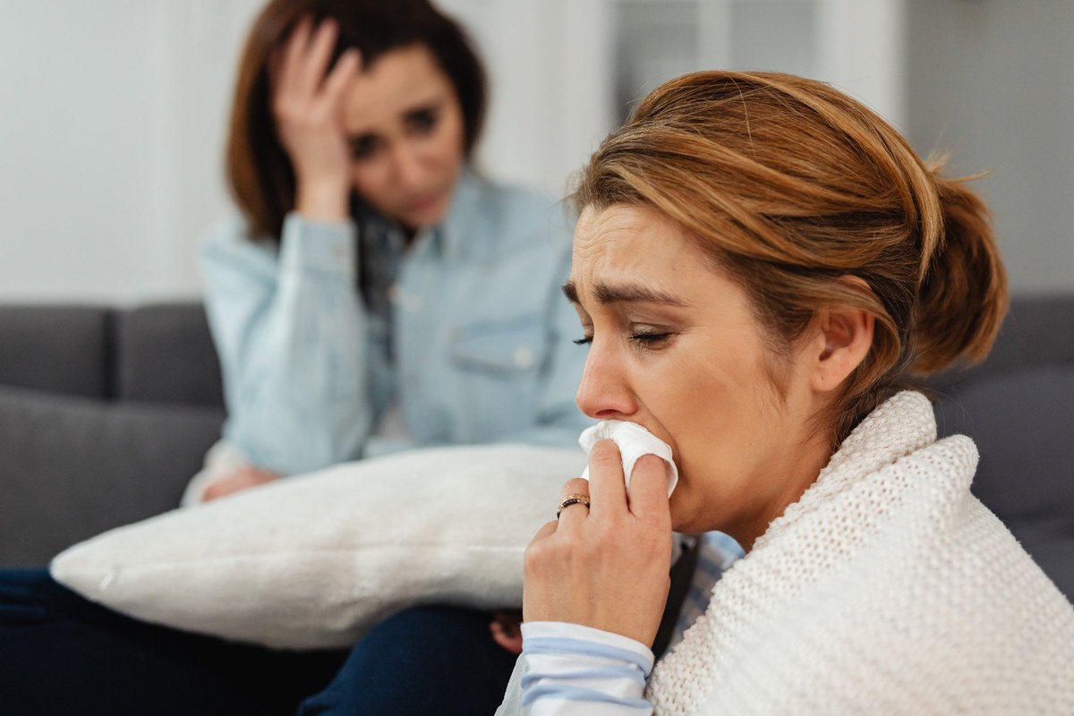 Woman crying into a tissue on a sofa while another woman looks on with worry